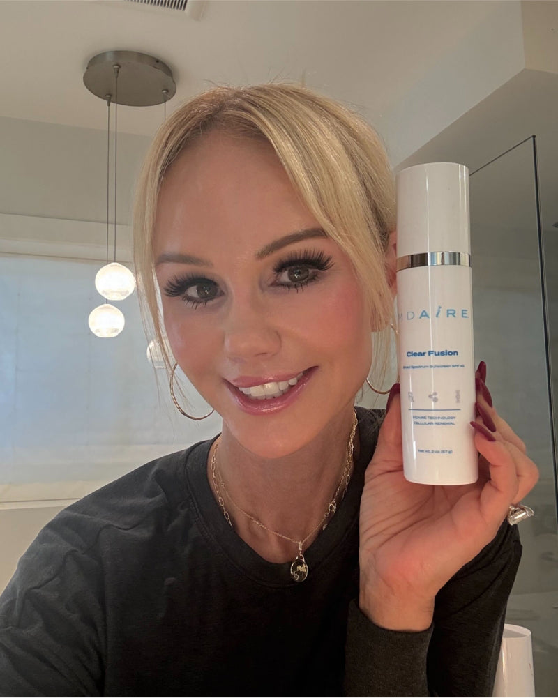 Woman holding Clear Fusion Sunscreen bottle with silver accents smiling in well-lit modern bathroom gallery position 1
