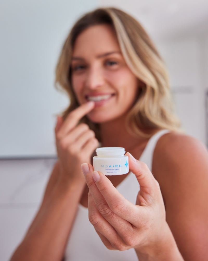 Woman holding open jar of Peptide Lip Mask close to lips with soft smile in gallery position 1