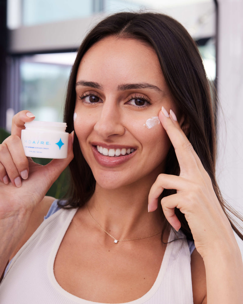 Woman smiling while applying NMF Intense Barrier Cream on cheek indoors in gallery position 1
