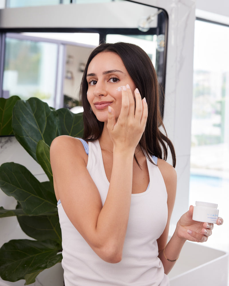 Woman applying NMF Intense Barrier Cream on cheek holding jar indoors in gallery position 1